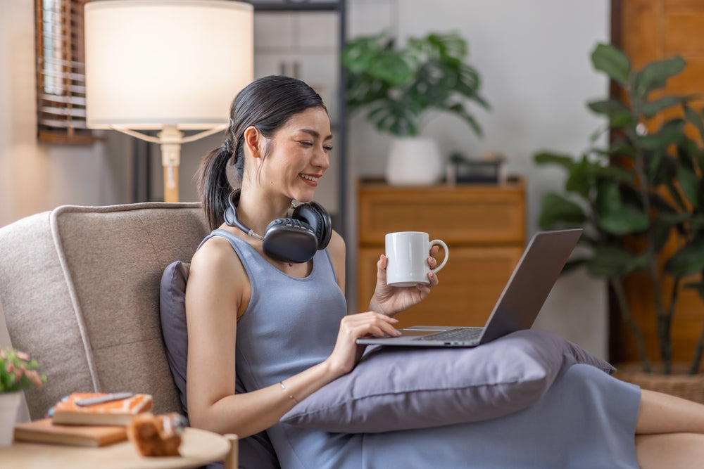 A woman sits on her computer with headphones on her neck and a cup of tea in her hand.